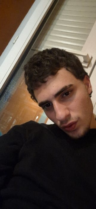 A light-skinned man with dark curly hair poses indoors. He looks directly at the camera with a slight tilt of his head. The background includes a window with closed white blinds. He wears a black shirt. The indoor setting appears to have a reflection on the window, adding another layer to the composition. The overall lighting appears to be soft and even.