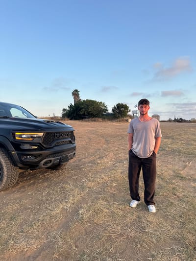 The image shows a young man standing in a field with a black Ram pickup truck parked nearby. The man is wearing a grey t-shirt and black pants. He is looking directly at the camera. His hands are in his pockets. He is standing on dry, brown grass. The pickup truck is parked on the same grass. The truck is facing the man. The truck's hood is open. The man and the truck are standing on a dirt path. There is a large tree behind them. The sky is blue with white clouds. The scene is located in the Bnei Zion neighborhood of Jerusalem, Israel, at coordinates (latitude: 32.226, longitude: 34.849). The image is taken from a slightly low angle. The lighting is bright and sunny. The image is likely taken in the late afternoon or early evening. The image is well-composed and has a natural, relaxed feel.