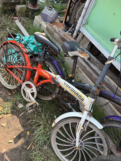 This image shows two bicycles and some debris, likely in a yard or outdoor storage area, given the presence of grass and dirt. One bicycle is white with yellow accents and features the text "YOKOHAMA" printed on the frame. It has a front wheel with a distinctive spoke pattern and a chainring. The other bicycle is primarily red with some black and purple elements. This bike has a visible chain, a rear rack, and is also identified as a "2.0 BIKE". The saddle of the white bicycle is torn, revealing the padding underneath. The scene is outdoors, with sunlight casting shadows, suggesting it is daytime. The weather appears to be clear. There is no specific activity or event taking place; the bicycles are stationary. The overall impression is one of disuse or storage, with the bicycles appearing somewhat weathered. The setting hints at a possibly rural or less maintained environment, consistent with the location context of Denasri Kulon, Indonesia. There is also a small wicker basket visible near the top left of the image.
