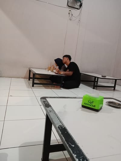A couple is seated at a low table in what appears to be a restaurant or food stall setting in Cangkring, Indonesia. The woman is wearing a hijab, while the man is in a black t-shirt and pants. They are sharing a meal and drinks. A green tissue box sits on a table in the foreground. The floor is tiled. A wall fan is visible.