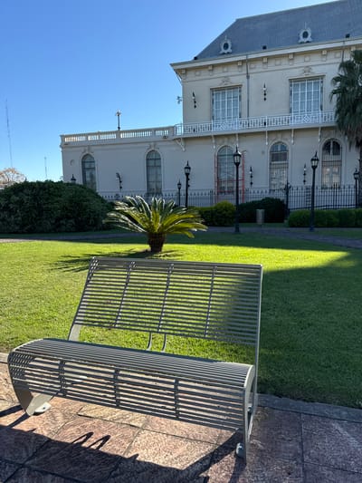 The image shows a metal bench on a stone pathway in Tigre, Argentina. Behind the bench is a green lawn, a small palm tree, and a section of a large, light-colored building with multiple windows and decorative elements. A black metal fence runs along the top of the building, and street lamps are visible in front of the building. The sky is blue. The scene is bathed in sunlight, creating strong shadows.