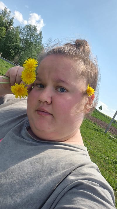 A woman is holding dandelions. One dandelion is in her hair. The location is Дойная, Russia. The background has grass and the sky. The woman is wearing a gray shirt.
