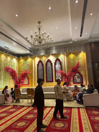 A gathering takes place indoors, possibly a wedding. The setting showcases a large chandelier hanging from the ceiling, which is adorned with numerous recessed lights. A backdrop of golden panels is ornamented with floral decorations and arch-shaped mirrors. People are seated on couches and chairs, and two men stand in the foreground, their profiles visible. The floor is covered by an ornate red carpet.