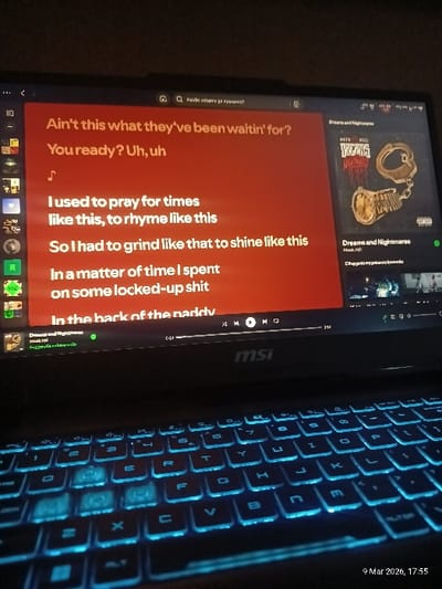 An MSI gaming laptop is depicted in a dimly lit indoor environment in Voiniagovo, Bulgaria. The laptop's screen displays song lyrics in white text on a prominent red background, showing lines such as "Ain't this what they've been waitin' for?" and "I used to pray for times like this, to rhyme like this." To the right of the lyrics, the album art for "Dreams and Nightmares" by Meek Mill is visible, featuring handcuffs and a pocket watch. Playback controls and a progress bar are at the bottom of the screen. The laptop's keyboard is illuminated with a bright cyan backlight. No individuals are present in the scene. A timestamp in the lower right corner indicates "9 Mar 2026, 17:55."