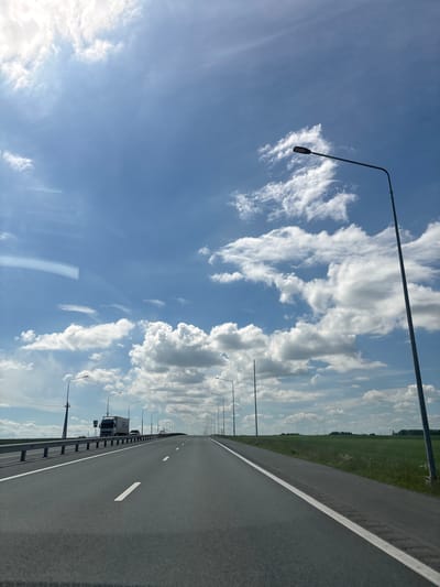 A wide asphalt road stretches into the distance under a bright blue sky filled with white, puffy clouds. The road has a white line marking the center and another on the right side. On the left side of the road, a metal guardrail runs along its edge. Several tall, grey lampposts are visible along the road, with one prominent lamppost dominating the right side of the frame. In the distance, a white truck and a car are traveling on the road. To the right of the road, a vibrant green field stretches out, meeting the horizon. The overall scene depicts a clear day on an open highway.