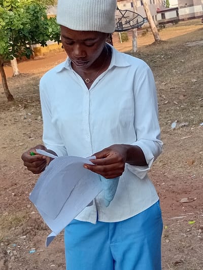 A person wears a white hat, white long-sleeved shirt, and blue pants, standing outdoors on a grassy area with some bare spots. The individual holds several white sheets of paper and a green pen, looking down at the documents. In the background, a building with a satellite dish is visible, along with a tree. The photo's orientation is rotated.