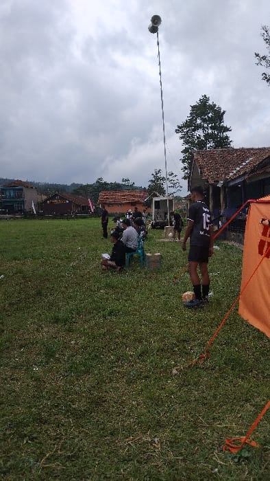 This image depicts an outdoor community gathering or event in Panjalu, Indonesia, under an overcast sky. The scene is set on a grassy field, with traditional houses featuring brown tiled roofs and lush trees visible in the background, hinting at a rural or village setting. Distant hills are also discernible on the horizon.
In the midground, a young man wearing a black and white sports jersey with the number "13" prominently displayed stands with a white football near his feet. He is positioned beside a partially visible orange canopy or tent structure, secured by orange ropes on the ground. Further into the midground, several people are seated on plastic chairs on the grass, with one individual in a grey shirt bending over, seemingly engaged in an activity like writing or examining something on the ground.
A tall pole equipped with two horn speakers near its top stands out on the left side of the frame, suggesting the presence of a public address system for announcements or music. The overall activity appears to be a sports-related event or a community function, possibly a local football game or a gathering involving youth, given the jersey and ball. The time of day is daylight, characterized by a cloudy, overcast sky, making it difficult to discern specific emotions, though participants seem engaged in the event.