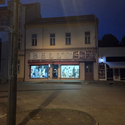 This image depicts a storefront in the city of Piešťany, Slovakia, at dusk or night. The primary focus is on a clothing store with the name "BELL'S" prominently displayed above a large display window. The window showcases mannequins dressed in light-colored clothing and accessories, illuminated from within. The building appears to be a multi-story structure with several windows on the upper floors. To the right of the clothing store, there is another storefront with a striped awning and a barber pole, indicating a different establishment. The street in front of the shops is paved, and there is a sidewalk area. A metal pole with some signage is visible on the left side of the frame. The overall lighting suggests it is evening, with artificial light illuminating the storefronts against the darkening sky.