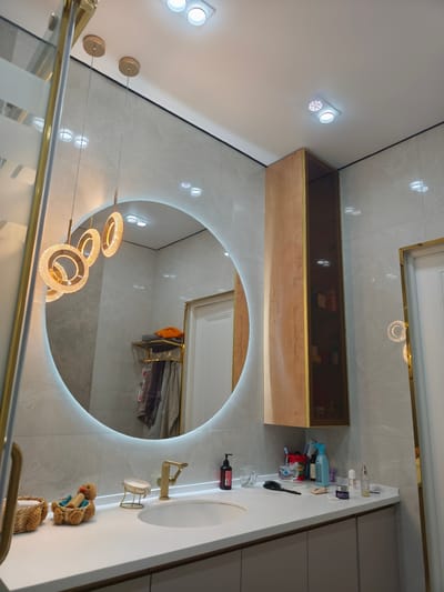 The image depicts a modern bathroom vanity area in Zhanaozen, Kazakhstan. A circular mirror with integrated LED lighting is the central feature, reflecting multiple decorative pendant lights and ceiling lights. To the right of the mirror, a tall, narrow cabinet with wooden front panels and glass shelves showcases various toiletries and decorative items. The countertop features a white sink with a gold faucet, a soap dish, a brush, and several bottles and containers of personal care products. On the left side of the vanity, two woven baskets are visible, one containing rolled towels and the other a small duck-shaped plush toy with a bar of soap. In the reflection of the mirror, a towel rack is visible, holding folded towels and other items. The walls are tiled with a light gray marbled pattern, and the ceiling is white with recessed lighting. A partial view of a glass shower enclosure with gold trim is present on the far left. A white door with gold trim is visible in the background reflection to the right. There are no people present in the image.
