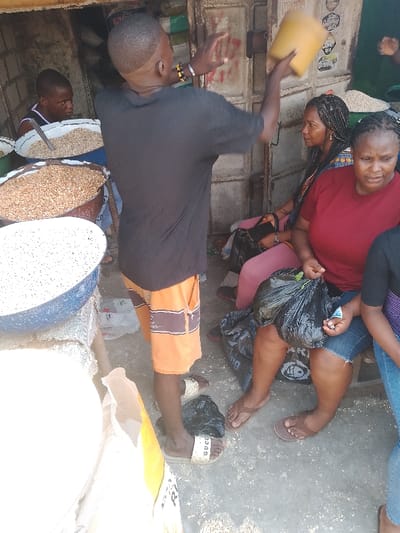 This image depicts a bustling market scene in Mgbuoshimini, Nigeria, under bright daylight conditions. The setting appears to be an outdoor or semi-outdoor stall, characterized by large metal containers filled with various grains and seeds, including what look like peanuts and lighter-colored cereals.

In the foreground, a man with a distinct haircut, dressed in a black t-shirt and orange and brown striped shorts, stands facing away from the viewer. He wears white "GUCCI" branded slides. His arms are raised, with his right hand holding a yellow plastic container aloft, suggesting he is in the process of measuring, scooping, or pouring goods.

To his immediate left, another man in a dark tank top is partially visible, leaning over a bowl of grains. On the right side of the frame, two women are seated, likely customers or fellow vendors. One woman, with braided hair and wearing a patterned top, looks towards the standing man. Beside her, another woman in a red t-shirt and blue jeans shorts holds a black plastic bag, indicating a purchase or merchandise. A larger black plastic bag rests on the ground near her, and another is visible near the standing man's feet.

The background shows a corrugated metal wall or door, typical of market structures. The ground is a simple concrete or dirt surface, with some scattered debris. The bright lighting suggests it is daytime, possibly mid-day. The overall scene captures the vibrant activity of a local market, with individuals engaged in commerce and daily routines. No prominent, readable text is visible other than the "GUCCI" logo on the slides.
