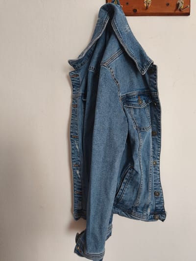 A blue denim jacket hangs on a wooden coat rack mounted on a textured off-white wall. The coat rack has four metal hooks and is affixed to the wall with screws. The jacket is the primary subject, occupying the majority of the frame. It is casually draped over the hooks, with its collar visible at the top and the sleeves and lower portion hanging down. The jacket has visible stitching, pockets with button closures, and metal buttons along the front. The background is a plain, lightly textured wall. The scene appears to be indoors, in a neutral setting. The location is Passignano sul Trasimeno, Italy.