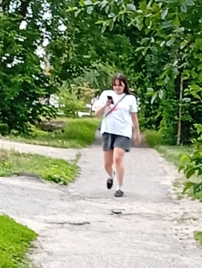 A young woman with dark hair, wearing a white t-shirt with a pink design and dark shorts, is walking on a dirt path. She is holding a smartphone in her right hand. The path is surrounded by green grass and trees. The scene appears to be a park or a residential area. The context provided places this image in Pavlovsky Posad, Russia.