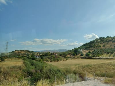 The image depicts a rural landscape in Raxa, Greece, under a clear blue sky with scattered white clouds. In the foreground, a dry, grassy field transitions into a verdant area with lush green foliage and some scattered purple flowers. A simple wire fence runs horizontally across the lower part of the frame.
Further into the scene, rolling hills dominate the midground and background. These hills are covered in a mixture of dry grasses, olive trees, and scattered bushes. A few white buildings with red-tiled roofs are visible nestled into the hillsides, suggesting a sparsely populated area. To the left, a tall, metallic electricity pylon stands prominently, indicating the presence of infrastructure. The overall impression is one of a sun-drenched countryside with signs of habitation and agricultural activity.