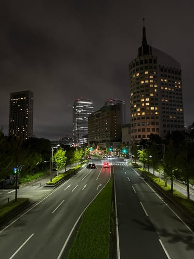 This is a nighttime, high-angle, long shot of a city street in Chiba, Japan, likely a business district, with several tall buildings and a road with moderate traffic. The sky is dark and cloudy, obscuring any visible stars. In the foreground, a two-lane road curves to the right, with white lane markings visible. Several cars are on the road, with one red car with its taillights on at the intersection in the midground, and another black car further up the road. Sidewalks are visible on either side of the road, lined with trees and manicured hedges. Several streetlights illuminate the scene.

Dominating the background are several skyscrapers. The tallest building on the right is a white, multi-story structure with a pointed spire on top. It has numerous windows, many of which are lit with a warm yellow light. To the left of this building is a similarly tall, slender skyscraper with a facade covered in horizontal bands of lights. Further to the left is a shorter, wider building with a beige facade and lit windows. Adjacent to the beige building is a light-colored, multi-story building with arched windows. In the far distance, other buildings are visible, some with illuminated windows, and what appears to be a red and white tower structure. The overall impression is one of a vibrant, modern city at night.