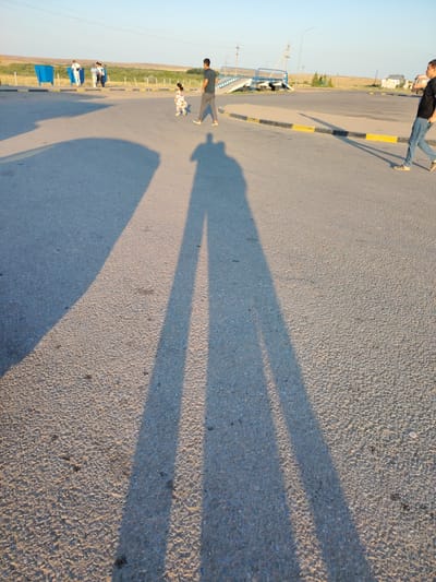 In Arshaly, Kazakhstan, a group of individuals are observed in an outdoor, open area with a paved surface. The scene features a young child in a floral dress walking towards a man wearing a dark t-shirt and grey pants. In the background, other individuals are visible near blue bins and a fence, with a bridge structure and utility poles also present. The ground is asphalt, marked with yellow and black lines. Long shadows of people are cast across the pavement, indicating a low sun angle. The environment appears arid with sparse vegetation and a clear sky.