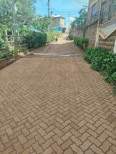 The image displays an uphill residential road in Kihara ward, Kenya, under a clear blue sky. The immediate foreground features a paved section constructed from brownish-grey interlocking concrete pavers arranged in a herringbone pattern. Approximately one-third into the scene, this paving transitions into an unpaved dirt road, with a small wet patch or puddle visible at the juncture.

On the left side, a wooden utility pole is visible near the paved section, adorned with several small notices. Behind it, a wooden fence precedes a dense line of green bushes and trees. Further in the background, a multi-story, light brown building and a structure with a blue roof are partially visible. Multiple utility poles with overhead power lines extend along this side of the road.

On the right side, low-growing green foliage lines the paved area. Beyond this, a concrete block wall, topped with security spikes or barbed wire, runs alongside the dirt road. This wall incorporates decorative stone cladding. A multi-story building with windows is situated behind this wall.

Three individuals are present on the dirt road, walking uphill. Two adults are observed together on the left side of the dirt path; one wears a yellow shirt. A third, smaller individual walks further ahead on the right side of the dirt path.