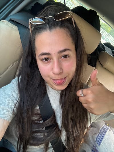 In Saransko, Bulgaria, a young woman with long, dark hair, wearing a light-colored sweater, is seated in the passenger seat of a vehicle. She is looking towards the camera, with her tongue sticking out and giving a thumbs-up gesture. Her sunglasses are perched on her head, and she is wearing two rings on her right hand. The interior of the car features beige leather seats and a black seatbelt. Outside the car, trees and buildings are visible. The lighting suggests it is daytime, and the scene is captured from a selfie perspective.