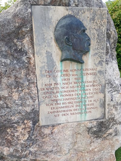 A weathered, grey stone monument is set against a large, rough, natural rock formation. The monument features a bronze bust of a man in profile, facing right, with a prominent mustache. Below the bust, red lettering is inscribed on a light-colored plaque. The text reads: "DER GEHEIME KOMMERZIENRAT DR. H.C. OTTO VON STEINBEIS (1839 - 1920) KAM 1863 NACH BRANNENBURG. ER MACHTE SICH ALS HOLZFACHMANN EINEN BESONDEREN NAMEN UND GALT ALS PIONIER DER DEUTSCHEN WIRTSCHAFTSGESCHICHTE. VON 1910 BIS 1912 ERBAUTE ER AUS EIGENMITTELN DIE ÄLTESTE ZAHNRADBAHN DEUTSCHLANDS AUF DEN WENDELSTEIN." The scene appears to be outdoors, with green foliage visible in the background.