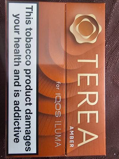 The image presents a close-up, eye-level view of a TEREA tobacco product box, designed for use with an IQOS ILUMA device, resting on a textured, dark brown surface. The box itself is a matte amber-brown color.
The top portion of the box features the brand name "TEREA" in large, light beige, embossed capital letters. To the left of "TEREA" is a distinct logo, composed of concentric, wavy, and irregular oval shapes in shades of light beige and amber. Below the main brand name, the word "AMBER" is visible in smaller capital letters, indicating the flavor or variant.
A horizontal line separates the upper and lower sections of the box. Centered below this line are the words "for IQOS ILUMA" in a light beige font, specifying the compatible device.
The most prominent feature of the bottom half of the box is a rectangular white warning label with a black border. This label contains bold black text stating: "This tobacco product damages your health and is addictive."
The background is a blurred, dark brown, textured surface, possibly fabric, emphasizing the product itself. The lighting is even, suggesting an indoor setting. There are no people or specific activities depicted. The clear English text on the packaging and warning label is consistent with the provided Holywell, United Kingdom location context.