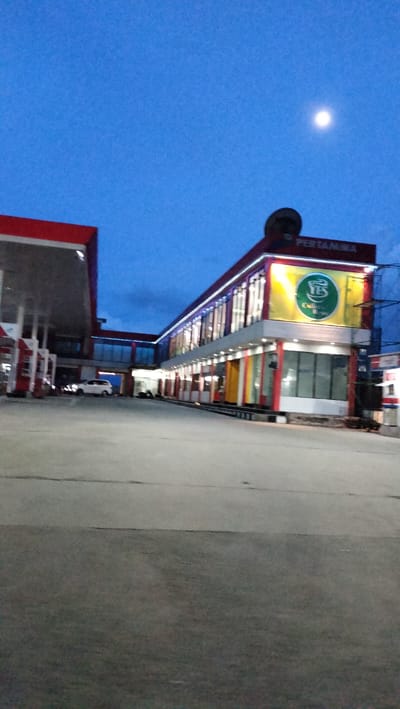 The image captures a **Pertamina** gas station and adjacent commercial building in **Kalalengkio, Indonesia**, during twilight. The **sky is a deep blue**, with a faint **full moon** visible. The gas station features a prominent red canopy over fuel pumps, with a white car parked nearby. The commercial building to the right has a modern design with a facade of large windows and colorful vertical panels. It is illuminated with interior lights, suggesting it is open for business. A large yellow banner with a green logo and the text "YES Coffee Tea" is prominently displayed on the building. The building is topped with a **Pertamina** sign. Scaffolding is visible on the right side of the commercial building, indicating ongoing construction or maintenance. The foreground consists of a paved area, likely a parking lot or driveway, with visible cracks and seams. The overall scene depicts a busy commercial hub in **Indonesia** at dusk, with the gas station and shops serving as focal points. The lighting suggests a transition from day to night, with the artificial lights of the buildings becoming more prominent.