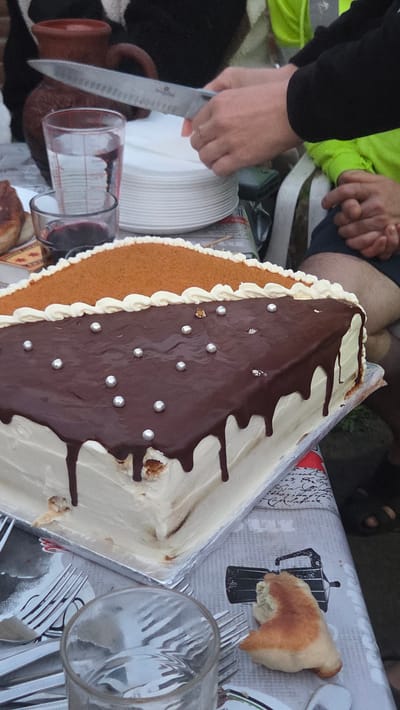 An overhead, medium shot captures a table setting for a dessert event. The focus is on a square-shaped cake with white frosting. The top of the cake is divided into two sections: one covered in a light brown, crumbly topping and the other with dark chocolate ganache drizzled over white frosting. Silver edible pearls are scattered across the chocolate section. A hand holding a knife is visible in the upper portion of the image, appearing to cut a slice of the cake. To the left, a glass filled with a red liquid sits next to a glass pitcher with a measurement scale. A stack of white plates is also present. The table is covered with a patterned tablecloth. The background shows glimpses of individuals and outdoor foliage, suggesting an outdoor gathering. The scene is located in the city of Killiney, Ireland.