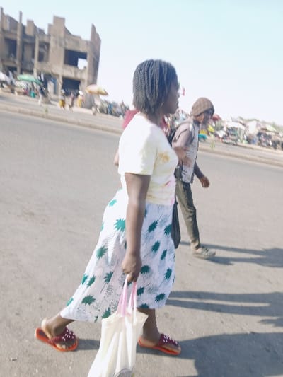 This image depicts an outdoor urban street scene in Kwekwe, Zimbabwe, under bright daylight. In the foreground, a dark-skinned individual with braided hair, wearing a light-colored t-shirt with faint stains, a white and green patterned skirt, and red slide sandals, walks from right to left across the frame. This individual carries a white tote bag with pink handles in their right hand. Behind them, slightly to the right, a second individual wearing a brown beanie, a light-colored vest over a dark shirt, dark trousers, and dark shoes, walks in the same direction. The background features a paved road bordered by a busy market area on the right, characterized by numerous stalls, people, and several open umbrellas, including one prominent yellow/orange and one green. To the far left, a large, multi-story concrete building, appearing unfinished or dilapidated, with exposed structural elements and empty window openings, stands under a clear, light blue sky. Strong shadows are visible on the ground, consistent with intense sunlight.