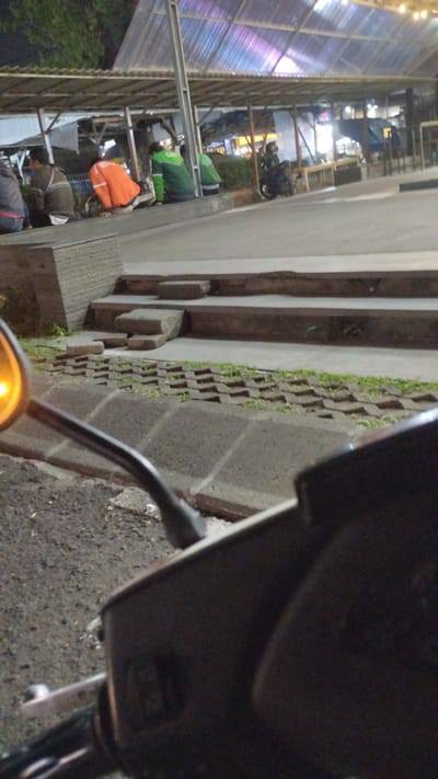 The image depicts an outdoor scene at night, likely near a food stall area or market, in Bringinbendo, Indonesia. In the foreground, the handlebars and part of a scooter are visible from a first-person perspective. The scooter's orange turn signal is illuminated. Behind the scooter, a paved walkway with decorative brick patterns is visible, leading to a series of concrete steps. Several individuals are seated along a low concrete barrier in the midground. One person wears a bright orange jacket. Others are dressed in green jackets, possibly indicating employment with a delivery service. A parked motorcycle is visible to the right of the seated individuals. The background features a covered structure with a corrugated roof, illuminated by artificial lighting that casts colorful reflections. The overall environment suggests a casual, possibly commercial, gathering space.