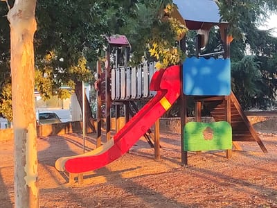 The image captures a playground with a red and yellow slide, a blue panel, and a green panel with a flower cutout. The playground structure is made of wood and metal and is situated on a ground covered with small stones. Trees with green foliage are visible in the background, and a tree trunk with rough bark stands to the left of the frame. The lighting suggests it is either early morning or late afternoon, with long shadows cast across the ground. In the background, a car is parked near a low stone wall. The scene appears to be in a public area, possibly a park or a schoolyard, located in Valverde de Leganés, Spain. There are no people visible in the image.