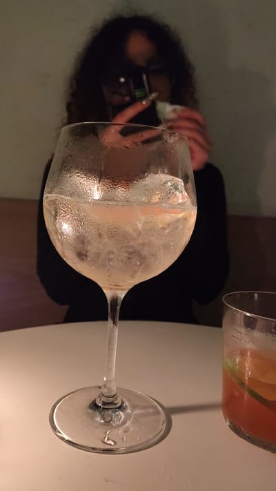 A rotated image presents a close-up of a large wine glass filled with ice, the glass is beaded with condensation. A second glass, containing a beverage with what appear to be fruit slices or garnishes, is visible in the top right corner of the frame. Behind the glasses, a person is visible using a phone, their dark curly hair and dark clothing contrast with the white table on which the glasses rest. The scene is dimly lit, and the location is potentially in the city of Barranco, Peru.