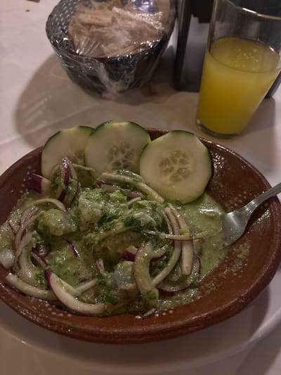 A still-life photograph shows a plate of ceviche in a terracotta-colored bowl alongside a glass of yellow liquid and a wrapped bundle, likely tortillas or bread, set on a white surface.
The ceviche consists of what appear to be shrimp or white fish mixed in a green sauce, garnished with sliced red onions and cucumber rounds. A fork rests in the ceviche. The terracotta bowl has a simple, rustic appearance.
The glass contains a yellow beverage, possibly juice or a mixed drink, and sits next to a dark object, perhaps a coaster or tray. The wrapped bundle is partially visible, encased in clear plastic. The white surface is likely a table. The overall composition suggests a casual meal setting, possibly in Veracruz, Mexico, given the prevalence of ceviche in the region. No people are visible in the frame.