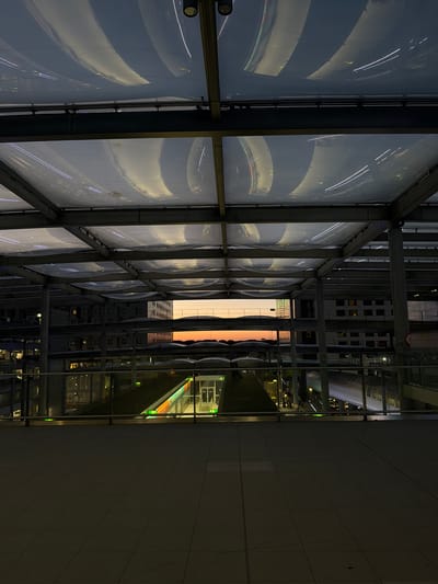 This image shows a view from a walkway structure at night, possibly a train station or airport terminal, in Crawley, United Kingdom. The structure has a metal frame and is covered by a translucent, wavy material that reflects lights from above. Below the walkway, there are several levels of buildings and infrastructure, with some windows illuminated. In the distance, a vibrant orange and yellow sunset is visible, contrasting with the dark surroundings. A train or similar vehicle is visible on a lower level, and there are people on a platform. The foreground is a dark, tiled floor.