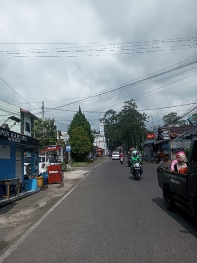 The image presents an overcast street scene in the city of Naung Bo Oe, Myanmar. An asphalt road, bordered by a white line on its left, recedes into the background. On the far right, the rear portion of a dark pickup truck is visible, displaying "deliver faster" text. Two individuals occupy the truck's bed: an adult male with a dark cap looking forward, and a female wearing a pink head covering. On the left, a blue building with "POS KAMLING RT 03 RW 05" inscribed on it features an attached wooden bench. Adjacent to this building are two red roadside fuel dispensers labeled "BENSIN," with a white SUV parked behind them. Multiple motorcycles are in motion on the road, with one rider in a green jacket and white helmet prominently featured driving towards the viewer. In the mid-distance, a tall, white mosque minaret rises, flanked by a conical evergreen tree on the left and various commercial structures and dense foliage on both sides of the road. Utility poles and a complex network of overhead power lines are visible against the heavily clouded, grey sky.