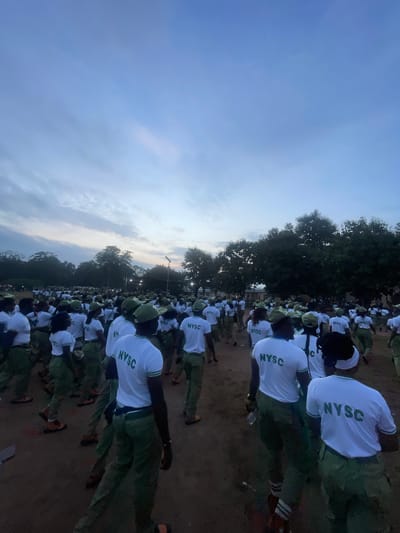This media file shows a large group of young adults, likely corps members, standing in formation outdoors. They are dressed in matching white t-shirts with the text "NYSC" printed in green on the back, and green trousers. The scene appears to be an open field or parade ground, with trees lining the background. The time of day seems to be early morning or late evening, indicated by the soft, diffused light and the color of the sky, which is a pale blue with some wispy clouds. The overall atmosphere suggests an organized event, possibly a morning assembly or drill, common during the National Youth Service Corps (NYSC) program in Nigeria. The individuals are facing in the same direction, indicating a collective activity. There are no overt displays of emotion, but the uniform attire and ordered arrangement suggest discipline and unity. The location context of Emure-Ekiti, Nigeria, places this scene within the cultural and administrative framework of the NYSC program in that region.