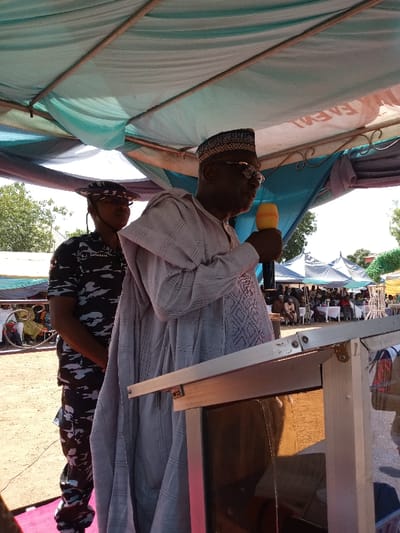 The image shows an older man wearing a grey robe and a hat, standing behind a wooden podium and speaking into a yellow microphone. He is wearing glasses. A younger man in camouflage fatigues is standing behind him, to the left. The scene takes place under a tent canopy with a blue and white patterned fabric. In the background, there are more people gathered under tents, as well as trees and a sandy ground. The location, determined from the given coordinates, is in the city of Zum, Nigeria.