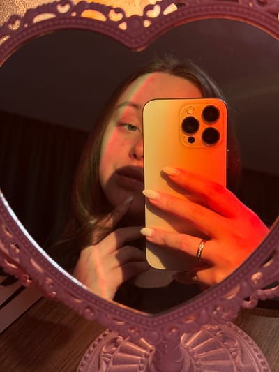 A person's reflection is captured in a heart-shaped mirror. The individual is holding a gold smartphone, obscuring part of their face. A hand with manicured nails and a ring is visible over the phone. The scene is bathed in warm, reddish-orange light, creating a soft, hazy effect. The mirror frame is ornate and pink.