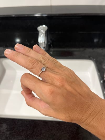 A close-up shot focuses on a person's left hand, adorned with a simple silver ring featuring a small, pale blue gem on the ring finger. The hand is positioned over a white sink, with a black countertop visible in the background. A chrome faucet is also present, slightly out of focus. The lighting suggests an indoor setting, likely a bathroom or kitchen. The image does not provide any contextual clues to suggest its location in Katunayake, Sri Lanka, or any specific environmental details beyond the immediate sink area.