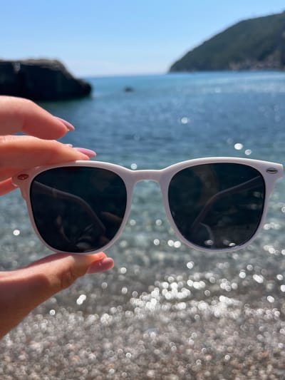 A pair of white sunglasses with dark lenses is held up by a person's hands. The sunglasses are in the foreground, and the background is out of focus, showing a body of water and a pebbly shore. The water is blue and appears calm, with sunlight reflecting off its surface. The shore is made up of small, light-colored pebbles. The scene suggests a sunny day, likely at a beach or seaside location. The person's hands have light pink painted fingernails and are holding the sunglasses by the temples. The overall mood is relaxed and summery. The location is identified as Zagrađe, Montenegro.