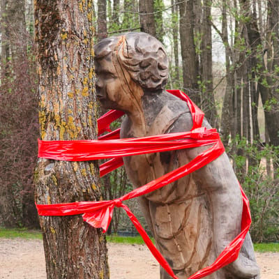 A wooden sculpture of a person is tied to a tree with red ribbon in an outdoor environment, likely a forest or park. The tree trunk is heavily textured with yellow-green lichen. The sculpture appears hand-carved and weathered. The red ribbon is wrapped around the tree and sculpture several times. The background includes other trees and foliage. The location is presumed to be Капцёўскі сельскі Савет, Belarus.
