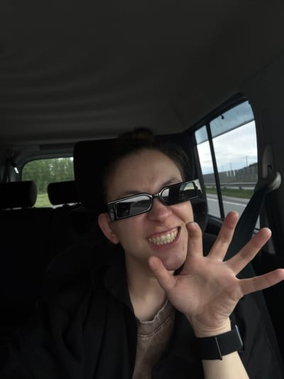 A woman is seated in a car with a sunroof. She has sunglasses tilted down on her nose, and her hand is raised with fingers spread. She is smiling. The car is moving on a highway. The location is Russkie Kirmeni, Russia. The environment shows the view through the sunroof, including a highway with infrastructure, and the car's interior. The woman is wearing a black top and has a watch on her left wrist.