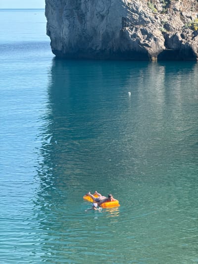 The image depicts a person relaxing on an inflatable raft in a body of water, likely a lake or sea. The person, wearing striped shorts, is lying on their back on a bright orange inflatable raft. The water is a clear, deep blue-green and appears calm with gentle ripples reflecting the sunlight. To the left of the frame, a rocky cliff face rises from the water, covered in sparse vegetation. The sunlight suggests it is daytime, with clear weather. The scene conveys a sense of tranquility and leisure, with the person enjoying a peaceful moment on the water. The reflection of the cliff and the sky on the water creates a striking visual contrast between the solid landmass and the fluid surface.