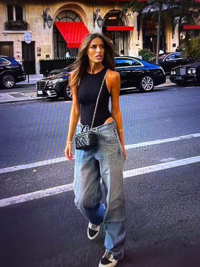 The media file shows a woman standing on a street in Beșalma, Moldova. She is wearing a black tank top, oversized blue jeans, and sneakers. She has a small black quilted purse with a silver chain strap. Her hair is long and brown, and she is looking at the camera. Behind her, there are several cars parked on the street. The buildings in the background appear to be shops or apartments with stone facades and red awnings. There is no visible text in the image. The lighting suggests it is daytime, and the weather appears to be clear. The overall scene depicts a casual street style moment.