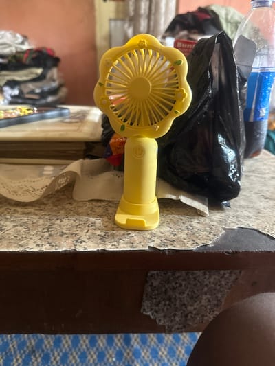 A yellow, flower-shaped portable electric fan stands upright on a speckled granite-patterned tabletop. The fan features a circular grill, visible blades, and a power button on its handle, which terminates in a small cradle-style stand. To the immediate right of the fan, a crumpled black plastic bag is present, with a clear plastic bottle containing a dark liquid visible behind it. The tabletop's front edge shows wear, exposing a darker material beneath the granite-patterned veneer. A white, possibly lace, fabric is partially visible beneath the fan. In the blurred background to the left, a pile of dark clothing is stacked, alongside flat objects. The far background reveals a solid, light reddish-brown wall. In the lower right foreground, a dark-skinned leg is partially visible. The floor beneath the table is covered with a blue and white checkered pattern. The scene is located within the city of Iriebe, Nigeria.