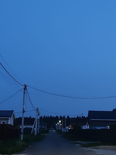 The image depicts a street scene in Skurygino, Russia, during dusk or early night. The sky is a deep, uniform blue. In the foreground and midground, utility poles with power lines are visible. On the left side of the road, a light-colored house with a dark roof and fence is partially visible. The road itself is paved and curves slightly to the right. On the right side of the road, a dark-colored house with a dark roof and white trim is prominent, with a bright light visible through one of its windows. Further down the road, streetlights are illuminated. A dense line of trees forms the background. The overall impression is of a quiet, residential area at twilight.