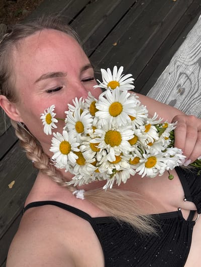 A person lies on a wooden surface, with a bouquet of daisies arranged around their head. The daisies are white with yellow centers, and some are placed directly on the person's face. The person's eyes are closed, and their expression is peaceful. Their hair is blonde and braided. They are wearing a black, sparkly bikini top. The wooden surface appears to be part of an outdoor seating area, possibly a bench or deck. The scene is set in Pozdyshki, Russia. The lighting suggests it could be daytime, with natural light illuminating the subject. The overall mood is serene and idyllic.