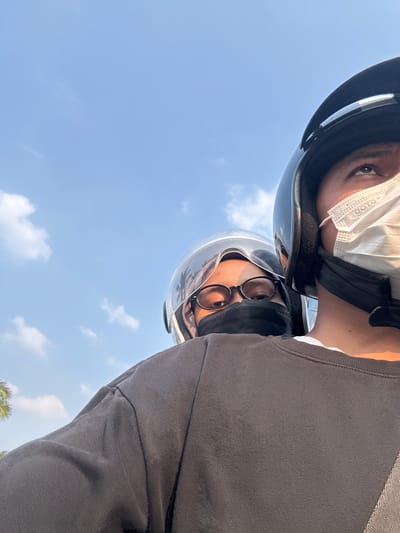 Two individuals wearing helmets and face masks are riding a motorcycle against a blue sky with scattered clouds, captured in a selfie-style shot. The person in the foreground, with a black helmet and a white mask, is looking upwards. Behind them, a second person with glasses and a black mask is visible. The scene appears to be in Pakulonan Barat, Indonesia, as indicated by the context, with a hint of trees visible on the left side.