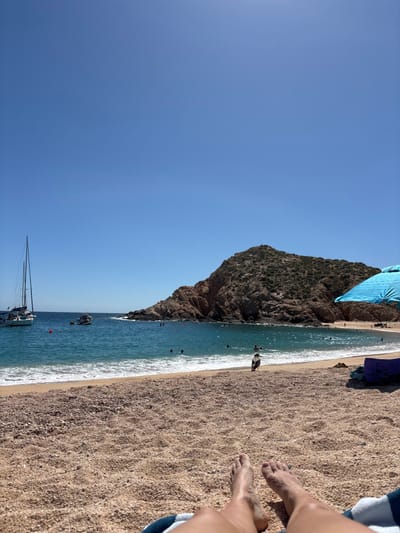 A sunny beach scene with a large rocky outcrop on one side and clear blue water meeting the sandy shore. Several boats and people are in the water and on the beach. A pair of feet is visible in the lower right corner on a towel. The clear blue sky takes up a significant portion of the image.