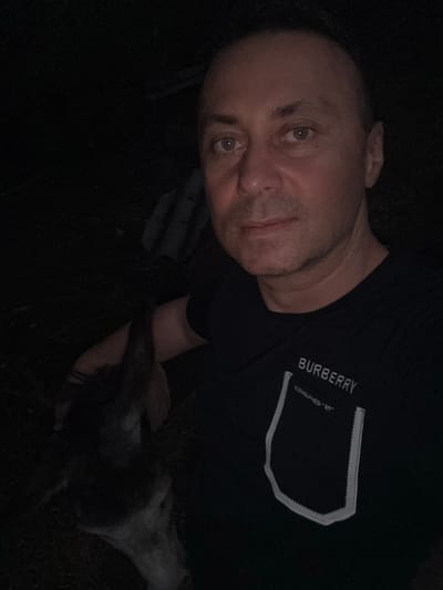 A selfie is taken by a man at night in an outdoor, dark environment. The man is wearing a black t-shirt with "BURBERRY" and "ESTABLISHED '85" printed on it. To the left of the man, a dark-colored animal's head is visible, appearing to be a dog. The background is mostly dark, suggesting it is night and possibly a rural or wooded area. There is no visible indication of the city of Tandzia, Georgia.