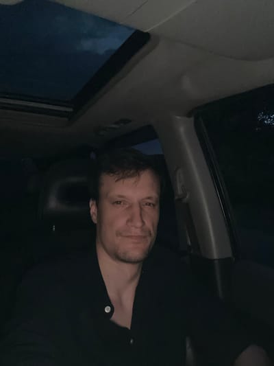The image shows a person sitting inside a car at night. The person is a man with short brown hair and is wearing a black collared shirt. He is looking towards the camera with a relaxed expression. The interior of the car is dark, with some ambient light coming from outside. A window on the left side of the car shows a dark, cloudy sky, suggesting nighttime or stormy weather. The image is taken from a slightly upward angle, focusing on the man's face and upper body. The overall mood is casual and intimate. There is no visible text in the image.