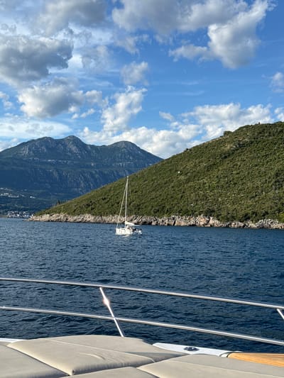 This image depicts a serene nautical scene in Luštica, Montenegro. A white sailboat with its sail up is gliding across a deep blue body of water, likely the Adriatic Sea, with people visible on its deck. In the foreground, the railing and seating area of a larger boat are visible, suggesting the photograph was taken from a private vessel. The background is dominated by lush green hillsides and rugged mountains, with a scattering of white buildings visible along the coastline. The sky is a vibrant blue, dotted with fluffy white clouds, indicating pleasant weather. The overall impression is one of tranquility and natural beauty.