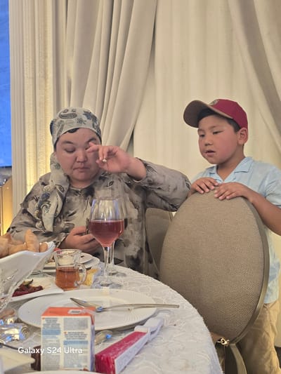 In Karakol City, Kyrgyzstan, a woman wearing a patterned headscarf and a printed shirt is seated at a table. She appears to be interacting with her eye, possibly with a finger. To her right, a young boy in a red baseball cap and a light blue shirt stands leaning on a chair. The table is set with plates, cutlery, a glass of red liquid, and a small glass of amber-colored liquid. There is also a white bowl containing food, and a small juice box is visible in the foreground. The background consists of draped curtains.